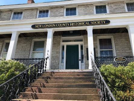 A view of the entrance to the New London County Historical Society, stone steps with wrought iron railings lead up to a porch with white columns and a stone historic house.