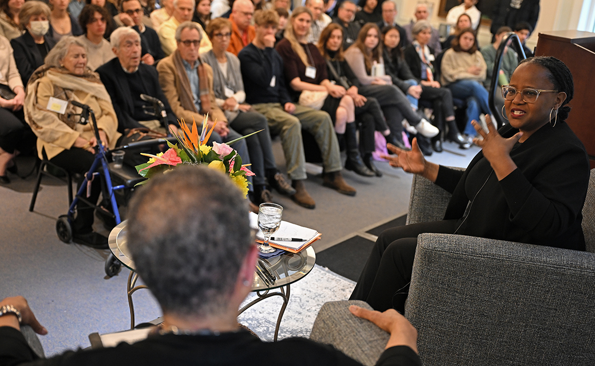 A woman poet speaks to an interviewer to her left in front of packed room of symposium attendees