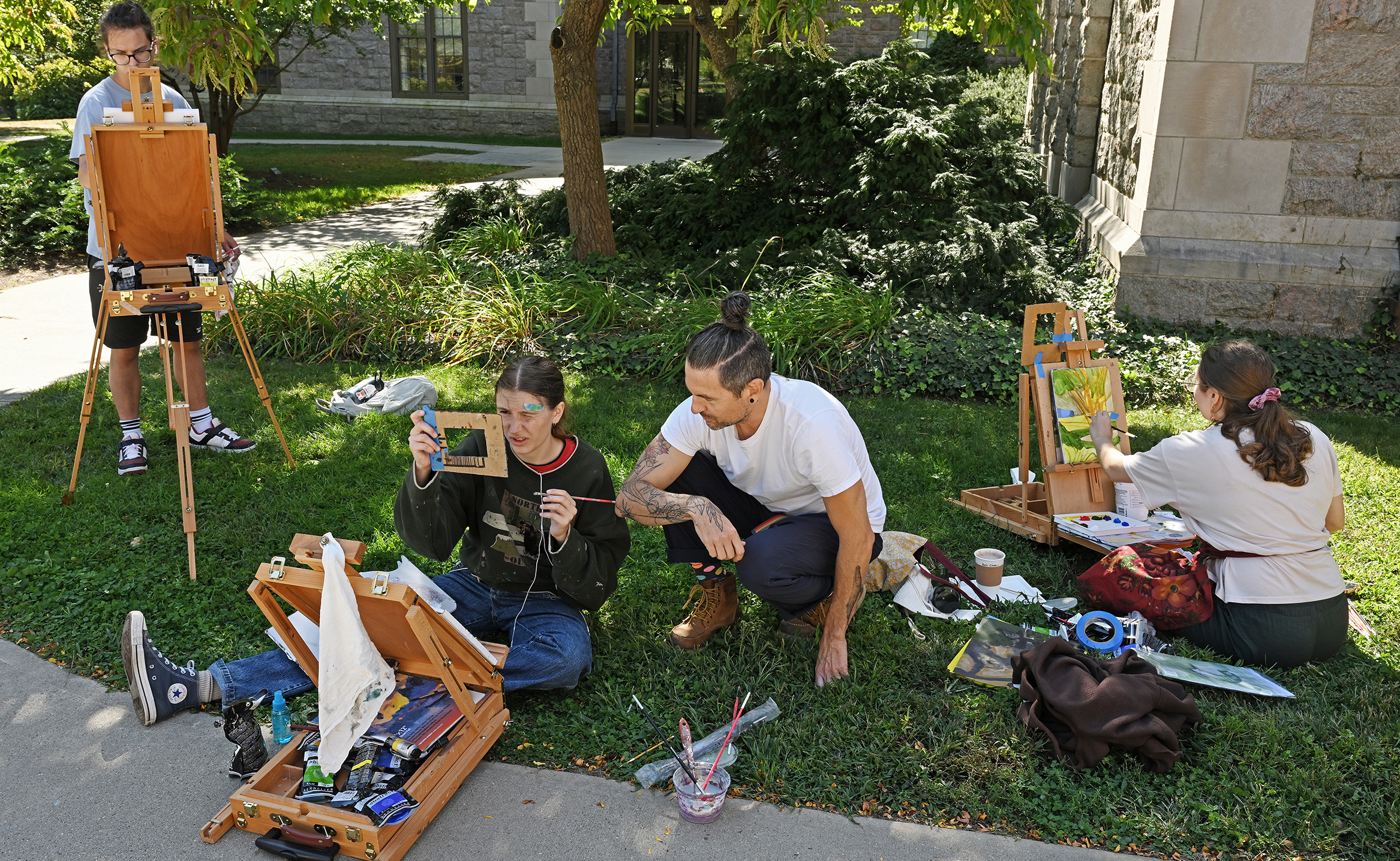 A professor in a white shirt mentors students painting outdoors.